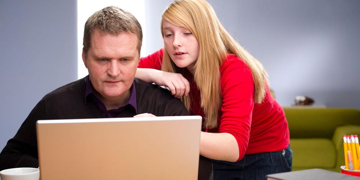 一个高中女孩从她父亲的身后望过去，而父亲正在用笔记本电脑