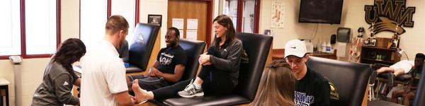 在运动训练设施里，萨曼莎·威尔逊教授坐在两名学生中间，一名学生裹着她的脚踝.